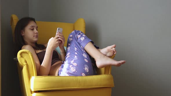 Young Female Sitting on Yellow Armchair Using Smartphone Mobile Device at Home