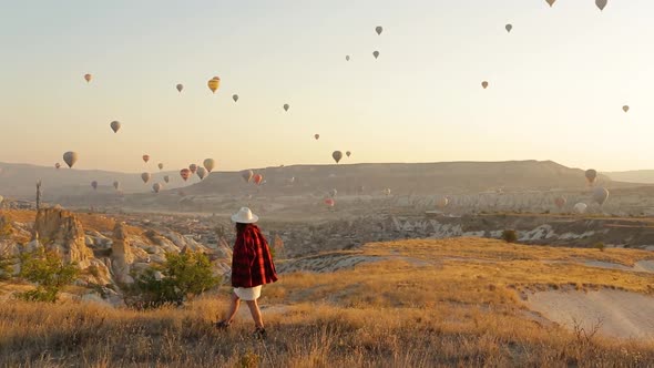 Woman In Hat Walking In Cappadocia. Colourful Hot Air Balloons Fly On Background