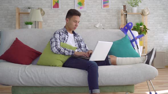 Asian Young Man with Broken Leg in Bandages Lying on Sofa with Laptop