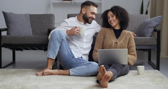 Young Family Multiethnic Couple Sitting Embracing on Living Room Floor Looking at Laptop Screen