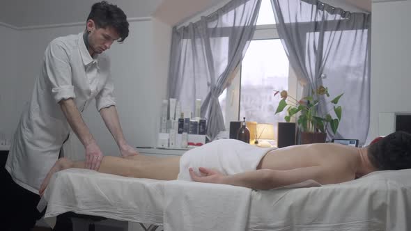 Wide Shot Side View of Concentrated Male Caucasian Masseur Massaging Legs of Relaxed Man Lying in