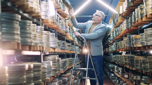 Archive Worker is Standing on a Ladder and Choosing a Film Tape