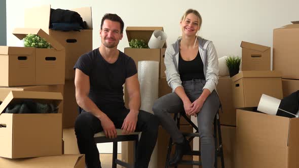 A Happy Moving Couple Sits on Chairs in an Empty Apartment and Nods at the Camera with a Smile