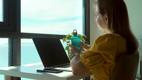 Smiling Woman Sitting on Table with Notebook and Drinking Fresh Cool Water