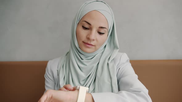 Cheerful Modern Muslimah Looking at Smart Watch Screen and Smiling Indoors in Cafe