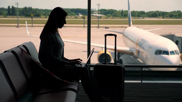 Dark Silhouette of Businesswoman with Luggage, Working on Laptop Against Panoramic Window, at