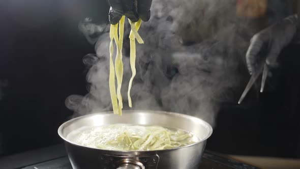 Chef in Black Gloves Putting Raw Pasta Into Boiling Water. White Smoke in Slow Motion Rising Above