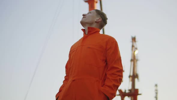 Handsome Young Container Warehouse Worker in Orange Uniform Standing By the Ship at the Harbor and