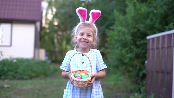 Little Blonde Girl with Bunny Ears Holding a Basket of Painted Easter Eggs in Her Hands and Laughing