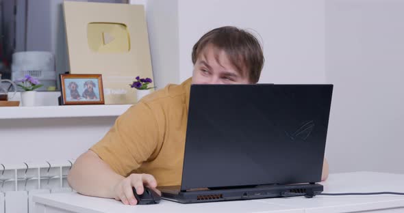 Young Man with Braces on His Teeth Sits Down at a Table Smiles Maliciously at Someone and Hides