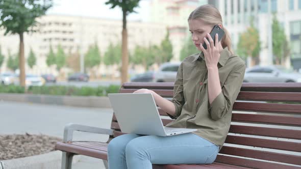Angry Woman Talking on Phone and using Laptop while Sitting Outdoor on Bench