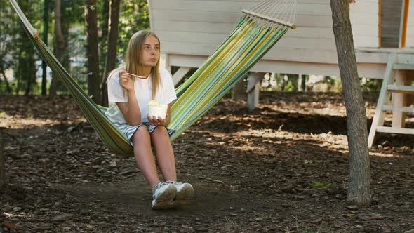 Young Female in Casual Clothes is Having a Rest Sitting in Multicolored Hammock and Eating Ice Cream