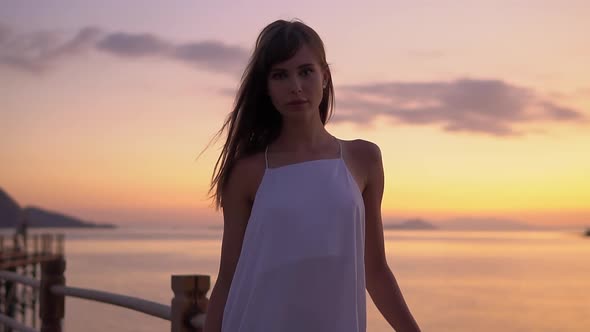 Portrait of Lovely Girl in White Dress Posing Outdoors with Sunset Sky in the Background Looking