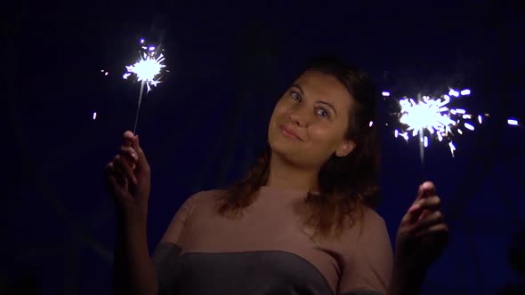 Beautiful Girl with Long Hair Holds Fireworks in Hands on a Background of Colorful Lights at Night