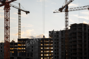 Cranes lift materials as workers construct a large building at a construction site illuminated by a