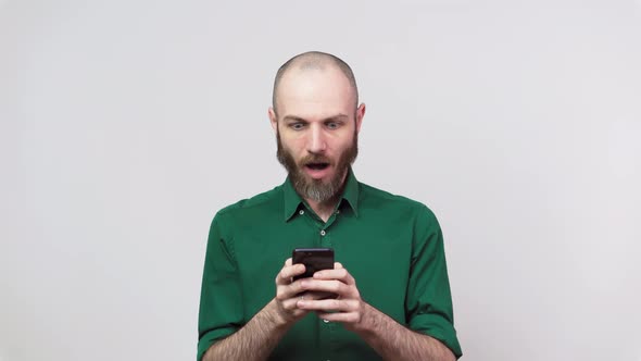 Handsome bearded man surprized and shocked using mobile phone over white background.