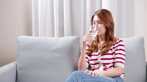 Asian young woman sitting on the sofa drinking water to relax during severe headache.