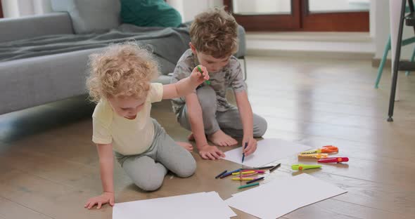 A Little Boy and a Little Girl are Drawing with Pencils on on the White Paper on the Floor Looking