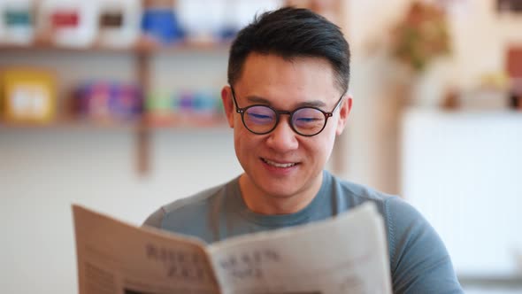 Cheerful Asian young man wearing eyeglasses reading newspaper