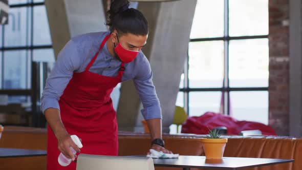 Mixed race male cafe worker wearing face mask disinfecting table