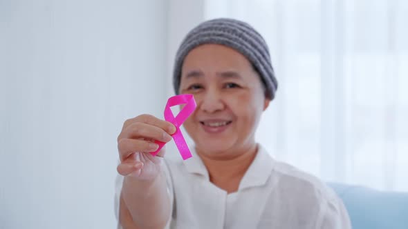 Elderly female patient holding pink ribbon in hospital. Breast cancer awareness concept.