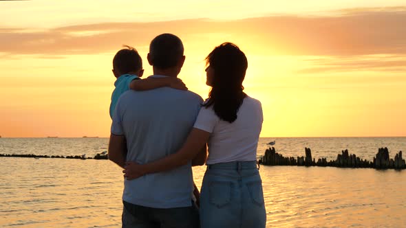 A Happy Family Enjoys the Sunset Over the Sea. A Father Holds His Son in His Arms. Mother and Son