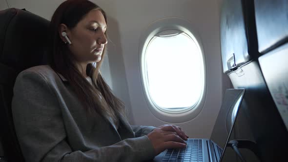 Brunette Woman in a Suit and a Wireless Earphone in Ear is Typing Text on a Laptop While Sitting on