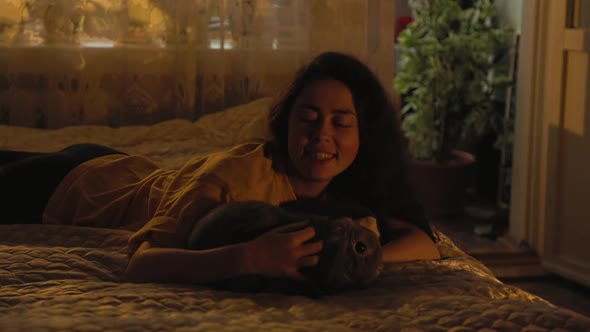 Smiling woman lying on her stomach on the bed and stroking a lop-eared gray rabbit. Pet care