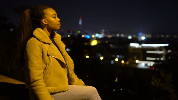 A Young Black Woman Looks Around and Smiles at the Camera As She Sits in an Urban Area at Night