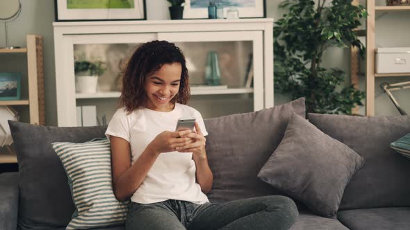 Beautiful African American Girl Is Using Smartphone and Laughing Relaxing on Comfortable Sofa at
