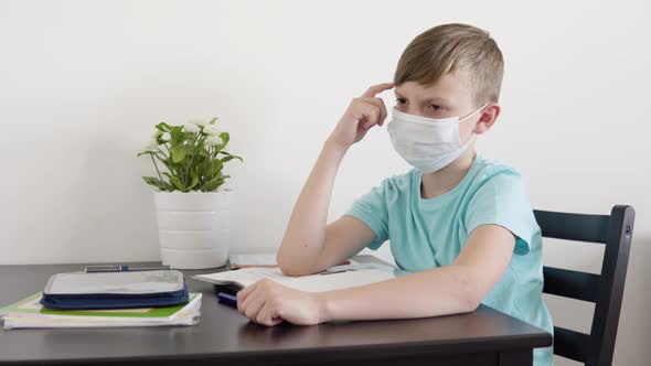 A Young Boy in a Face Mask Thinks Hard As He Does Homework for School at a Table at Home