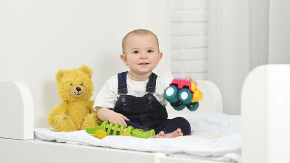 a Baby Boy in a Denim Jumpsuit Smiles and Plays with Toys While Sitting on a Bed