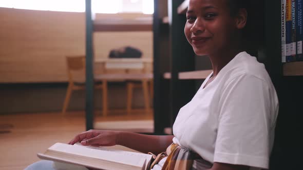 A schoolgirl sitting in front of library's bookshelf and reading a book preparing for an exam.