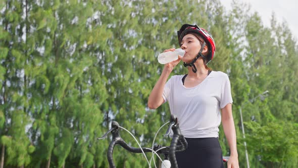 Asian woman take a break, drink water from bottle after riding bicycle in the evening in public park