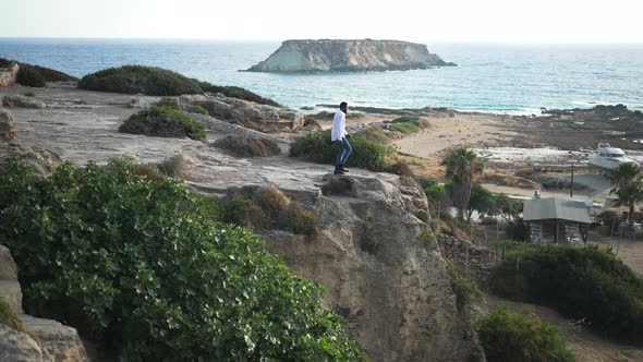 Extreme Wide Shot of African American Male Tourist Stretching Hands Standing on Cliffs on Blue