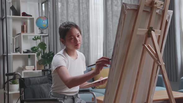 Side View Of Artist Girl In Wheelchair Holding Paintbrush Thinking And Painting On The Canvas