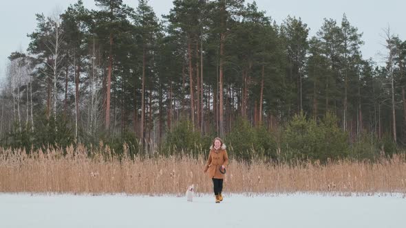 A girl in a coat and yellow boots runs in winter on the ice of the lake with a dog in clothes