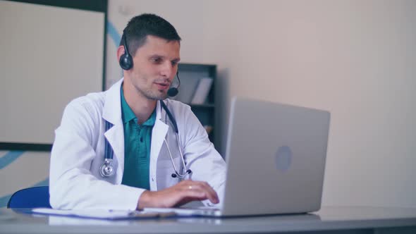Friendly Male Doctor in White Medical Coat With Headphones Making Conference Call on Laptop