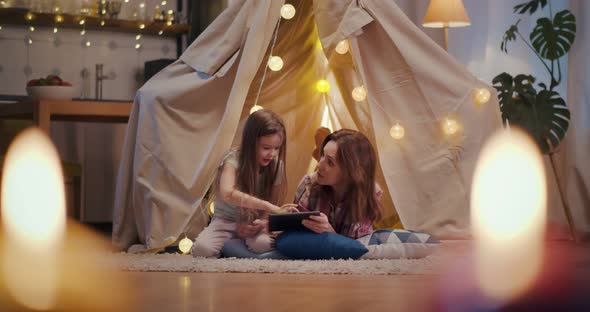 Mother and Little Daughter with Tablet Computer Playing in Teepee at Home in Evening