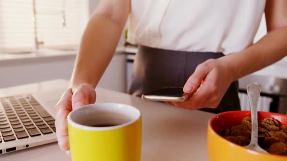 Woman using mobile phone and laptop while having coffee in kitchen