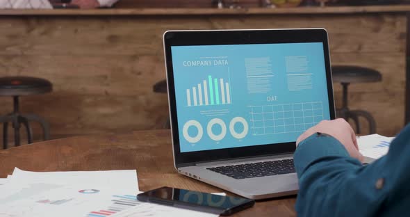 Man Sits at a Coffee Shop Table and Looks at the Company Data Graphs and Diagrams