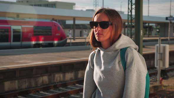 Portrait of young brunette woman in sunglasses and backpack waiting for train at railway station