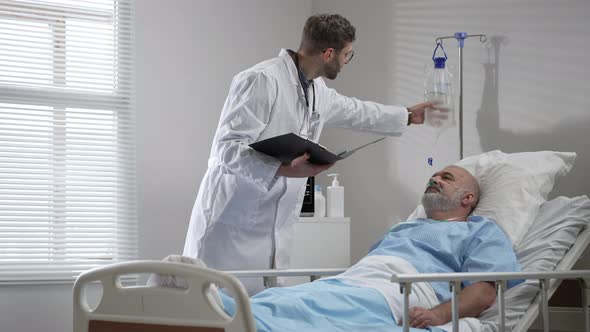 Young Doctor in Glasses Talking to Senior Man Resting in Hospital Bed