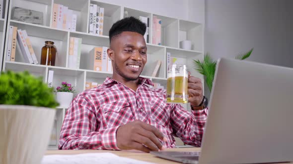 African man working from home taking vdo call on laptop with colleague and drinking beer 