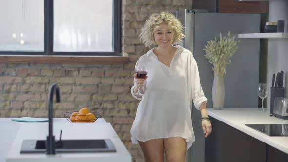 Gorgeous Tattooed Slim Woman Walking with Wine Glass in Kitchen Dancing and Smiling