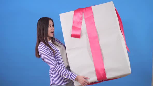 Positive Young Asian Woman in a Shirt Holds a Huge Gift in a Box on a Blue Background