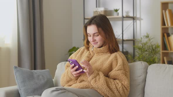 Attractive Teenage Girl Sitting on the Sofa at Home Using Phone and Smiling