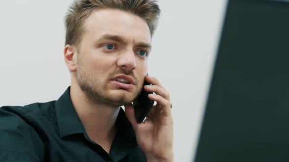 Handsome Young Bearded Man Talking on Mobile Phone and Using Laptop at Office.