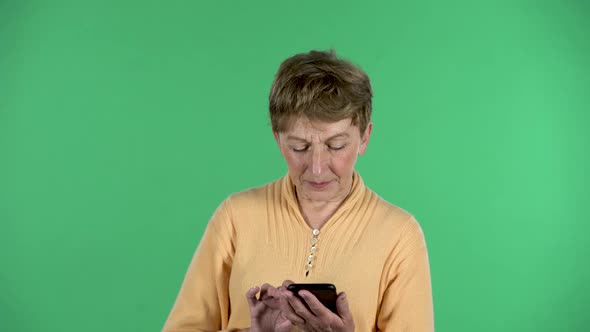 Portrait of Elderly Woman Is Texting on Her Phone Isolated Over Green Background.