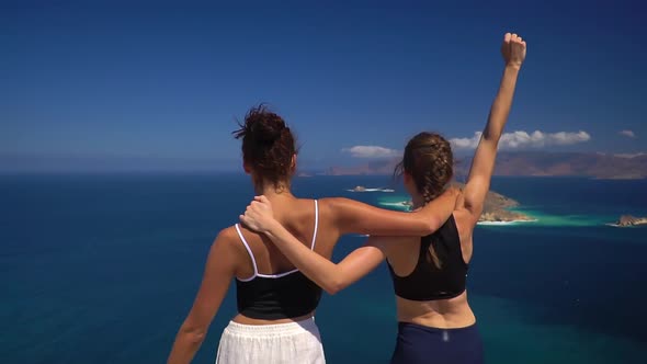 Two Athletic Brunette Girls Rejoice View From Top of the Hill in Sunny Day with Ocean on Background